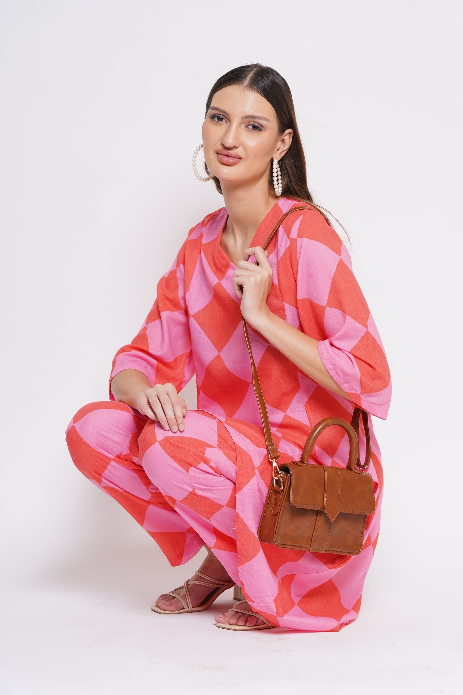 A woman crouches in a studio wearing a bright pink and coral geometric print kurta and matching trousers, accessorized with a brown leather shoulder bag, large hoop earrings, and flat sandals, against a plain light gray background.