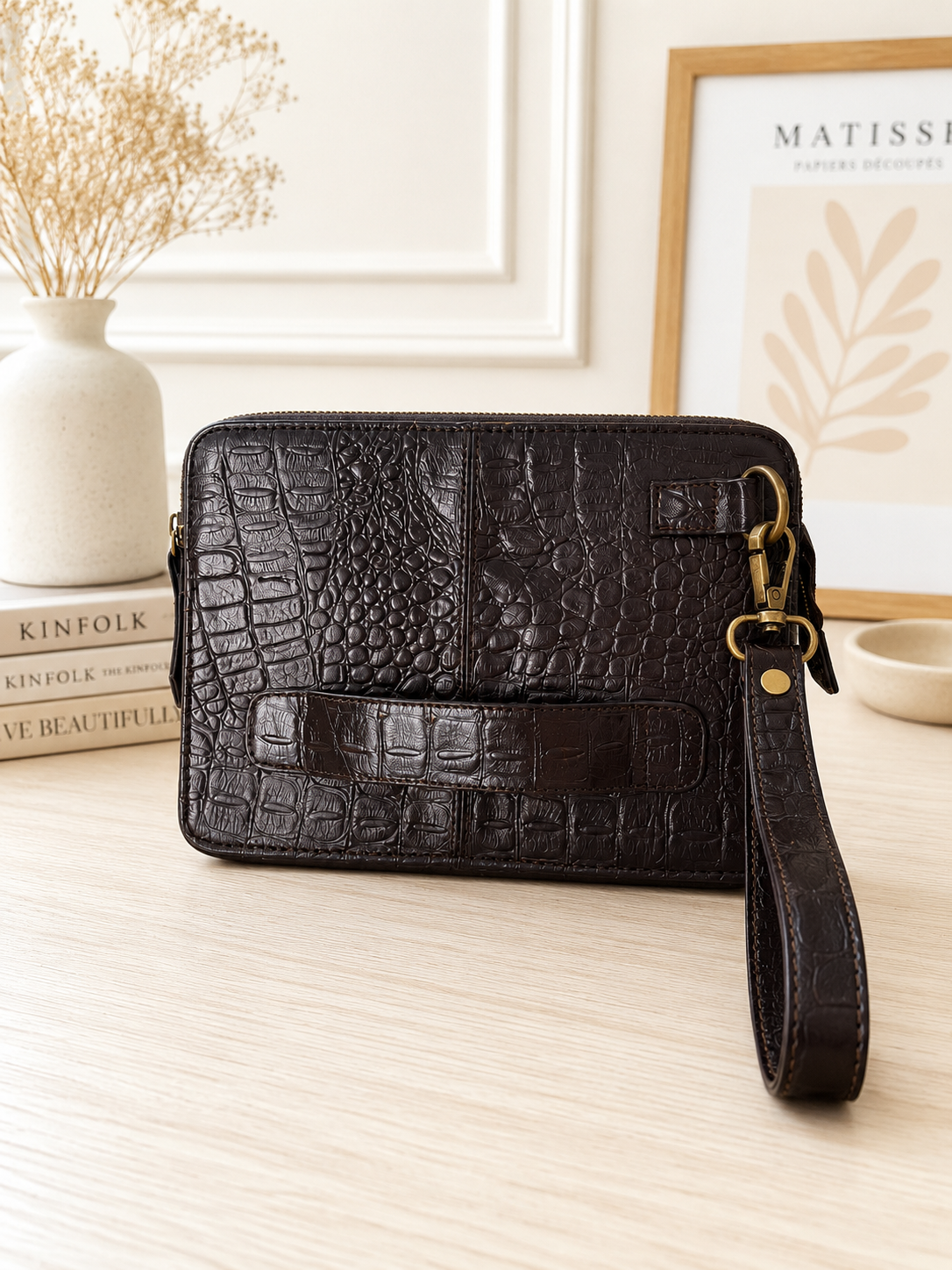 A dark brown crocodile-embossed leather wristlet clutch with a matching wrist strap and gold-tone hardware, displayed on a light wood tabletop with decorative books, a vase, and framed wall art in the background.