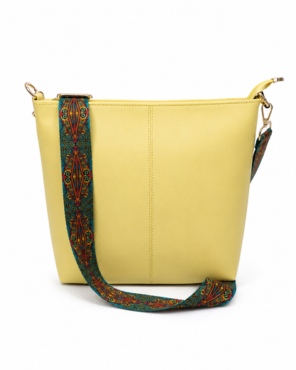 A light yellow faux-leather shoulder bag with a wide, patterned green fabric strap featuring red, orange, and yellow geometric designs, shown on a white background.