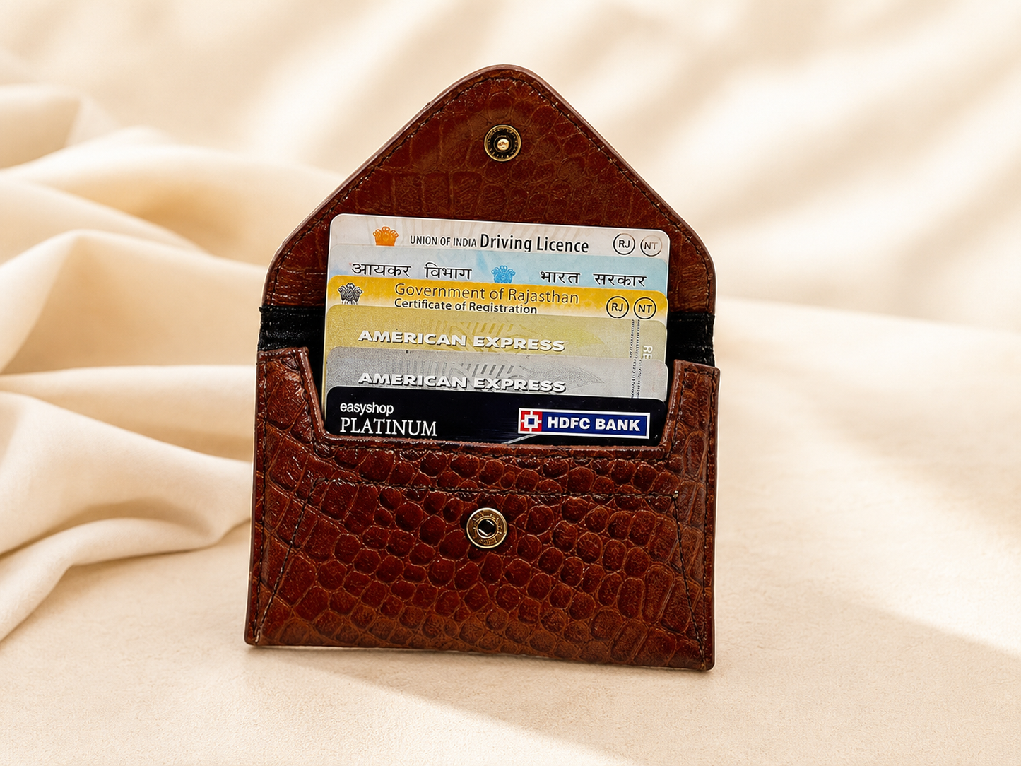 A brown crocodile-embossed leather card holder wallet with a small top button, shown open with sample credit and debit cards inserted, set against a draped beige fabric background.