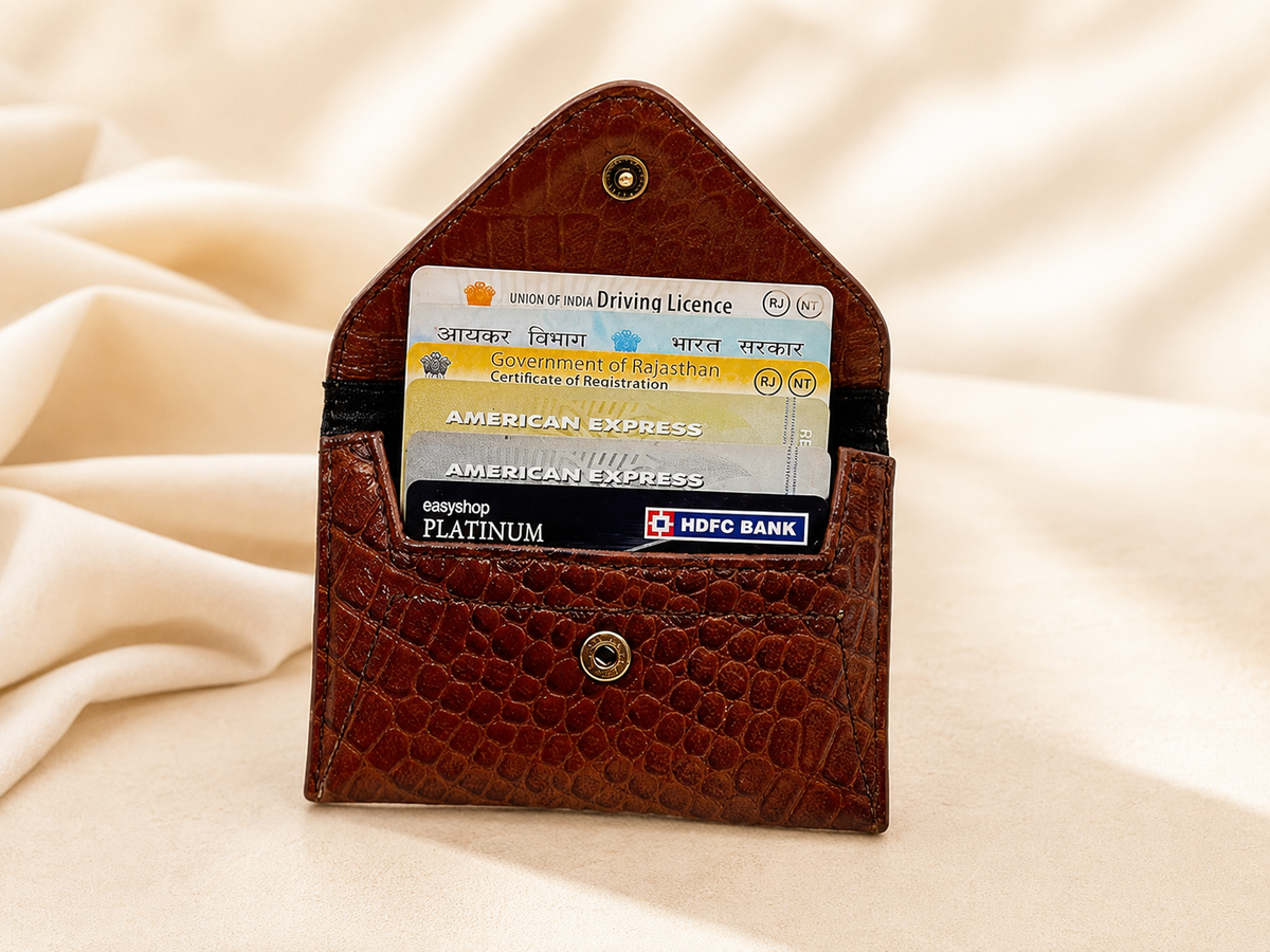 A brown crocodile-embossed leather card holder wallet with a small top button, shown open with sample credit and debit cards inserted, set against a draped beige fabric background.