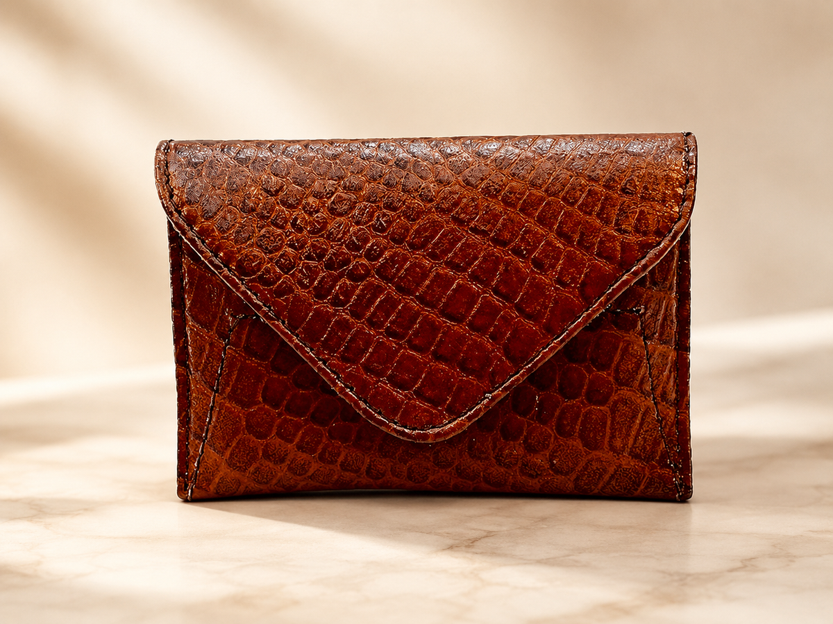 A brown crocodile-embossed leather envelope wallet standing upright on a light marble surface against a warm beige background.