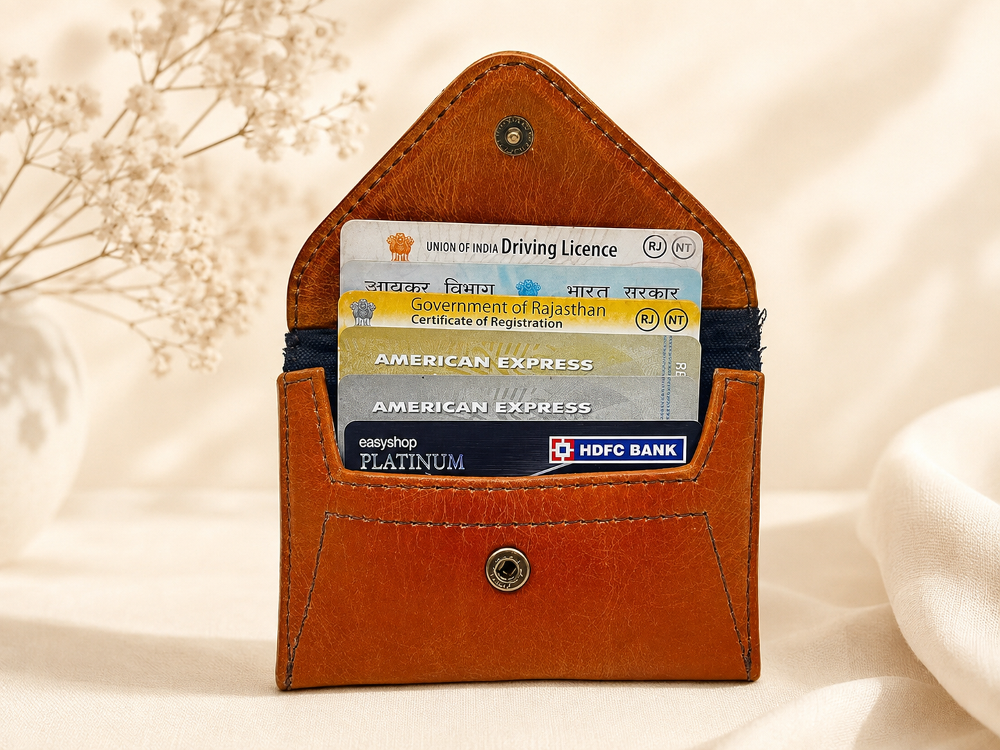 A brown leather card holder wallet with a pointed top and snap button closure, shown holding several cards including a Union of India Driving Licence, American Express, and HDFC Bank. The wallet is photographed on a soft beige fabric background with dried flowers and a white vase as styled props.