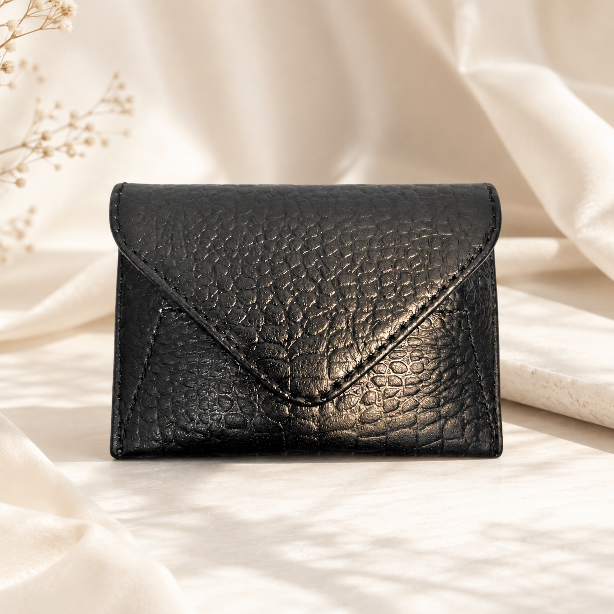A black textured leather envelope-style wallet or card holder, photographed on draped cream fabric with soft natural shadows.