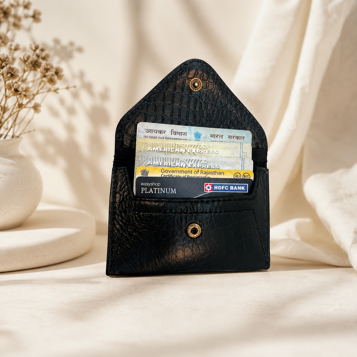 A black textured leather card holder wallet standing upright on a draped cream fabric surface, with two sample cards inserted into its slots. A small gold-tone snap button is visible at the bottom, and dried flowers in a white vase sit to the left.