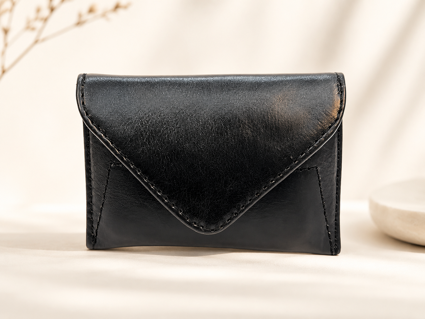 A small black leather envelope-style card holder or wallet with visible stitching, photographed on a warm off-white surface with soft shadows and a decorative dried branch in the background.