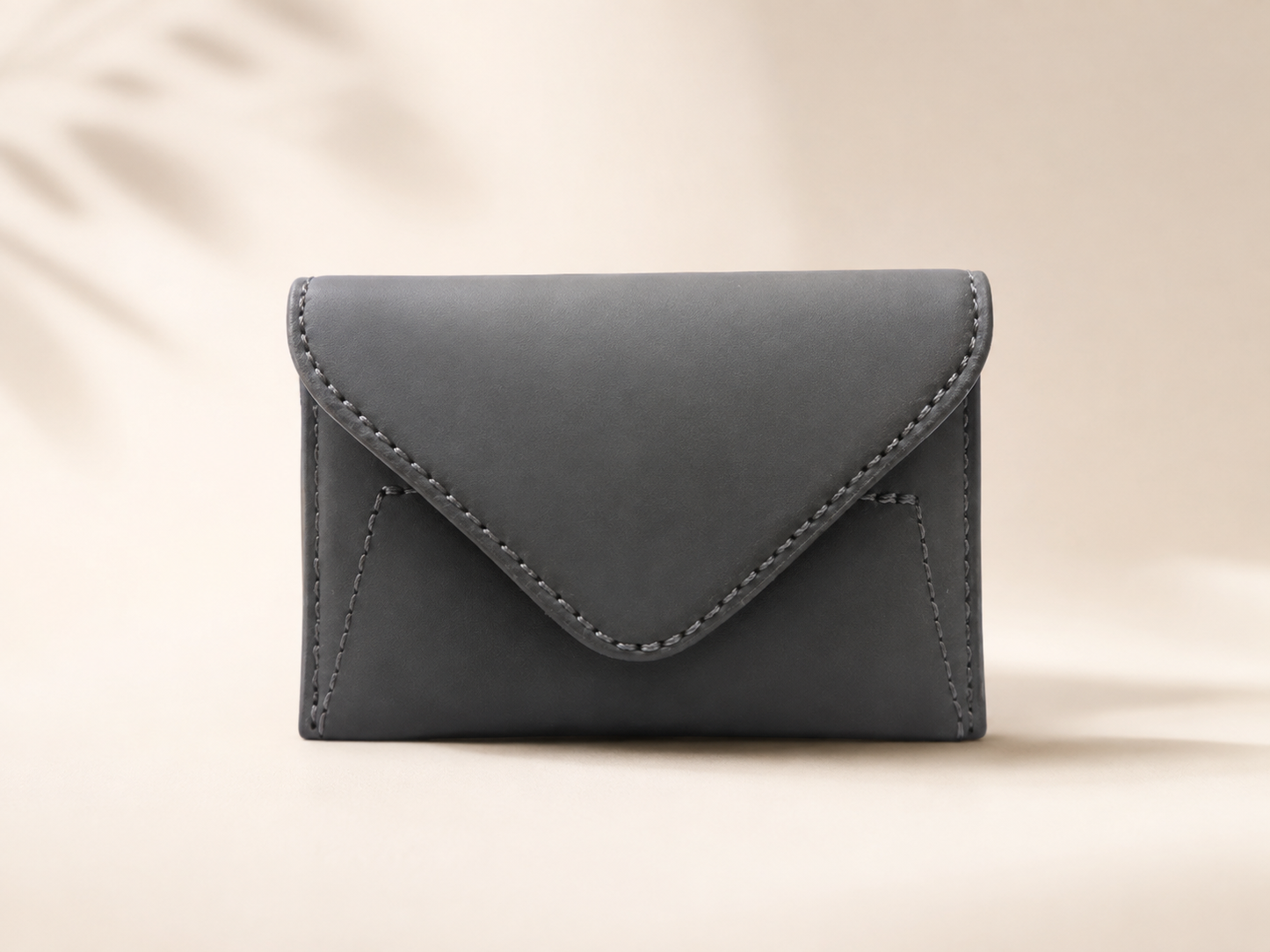 A dark gray envelope-style card holder or small wallet with visible stitching, centered on a warm beige background with soft leaf shadows.