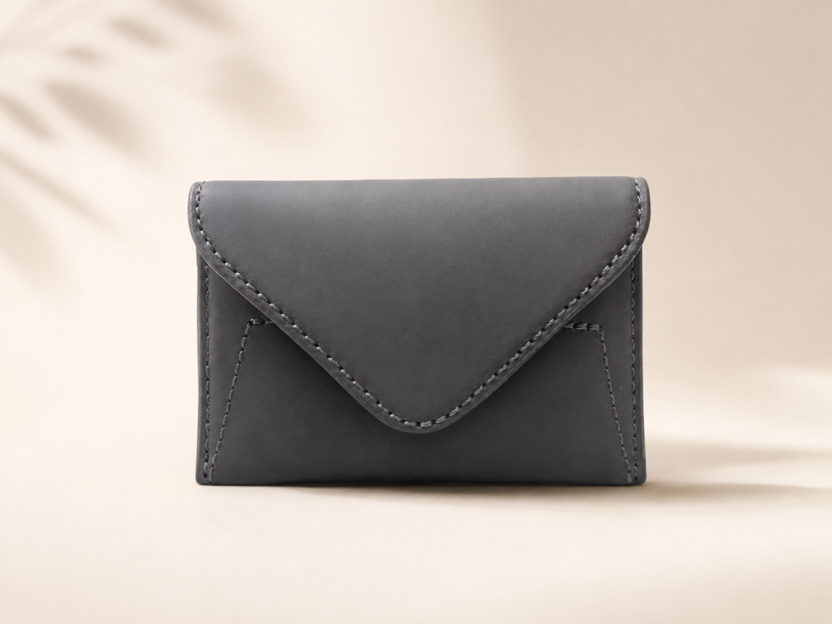A dark gray envelope-style card holder or small wallet with visible stitching, centered on a warm beige background with soft leaf shadows.