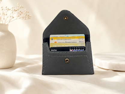A dark gray leather card holder wallet with brass snap buttons, shown open with two cards inserted. The wallet is styled on a draped cream fabric surface with a white ceramic vase and dried flowers on the left and a white marble slab on the right.
