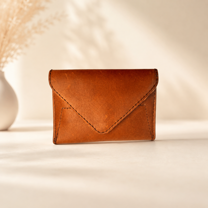A small brown leather envelope-style card holder wallet standing upright on a light beige surface with soft shadows and a blurred decorative vase in the background.