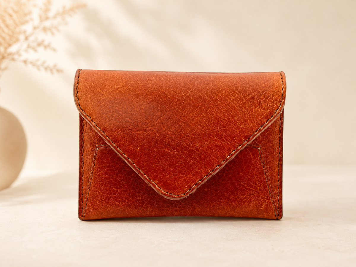 A small brown leather envelope-style wallet with visible stitching, photographed upright on a light beige surface against a soft cream background with a blurred decorative vase and dried stems at the side.