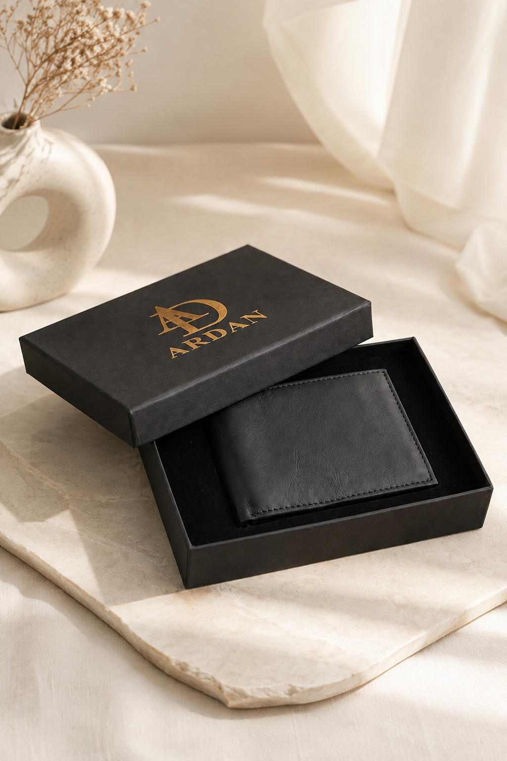 A black leather wallet presented in an open black gift box with gold 'ARDAN' branding, styled on a light beige surface with soft draped fabric and dried flowers in the background.