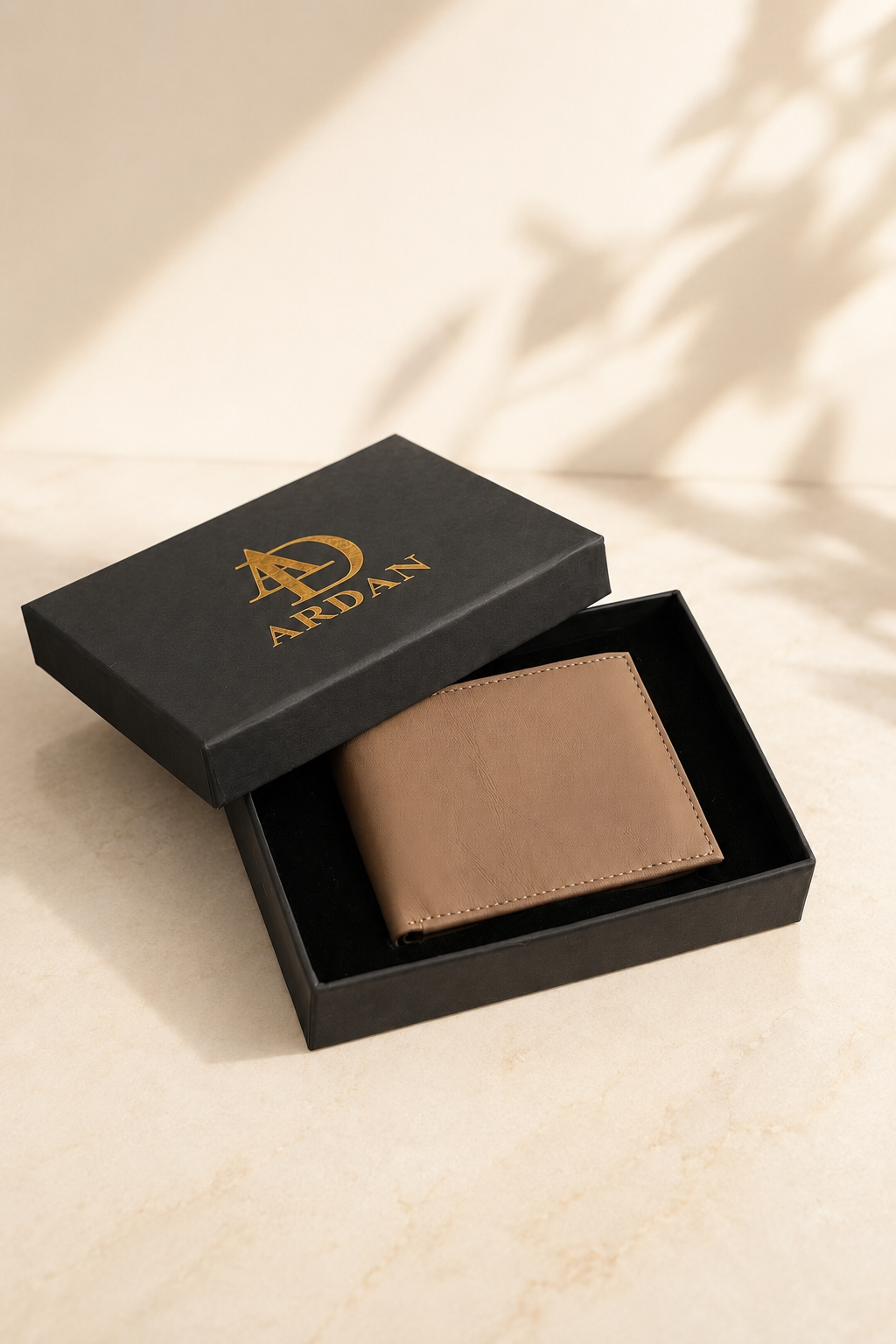 A tan leather bifold wallet presented in an open black gift box with the lid beside it, both branded with a gold ARDAN logo. The scene is styled on a light stone surface with soft leaf shadows.