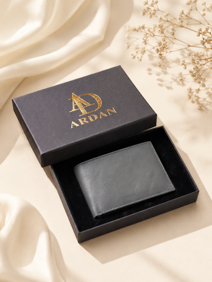 A black leather bifold wallet presented in an open black gift box with a gold-embossed ARDAN logo, styled on a cream satin fabric with dried floral accents.