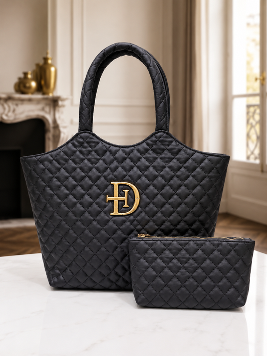 A black quilted tote bag with a large gold 'HD' monogram logo on the front, displayed alongside a matching black quilted pouch on a white marble surface in an elegant interior setting.