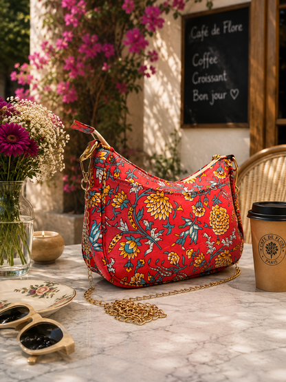 A pink shoulder bag with a colorful floral print and a gold chain strap, styled on a marble table in a cafe setting with a coffee cup, sunglasses, and flowers.