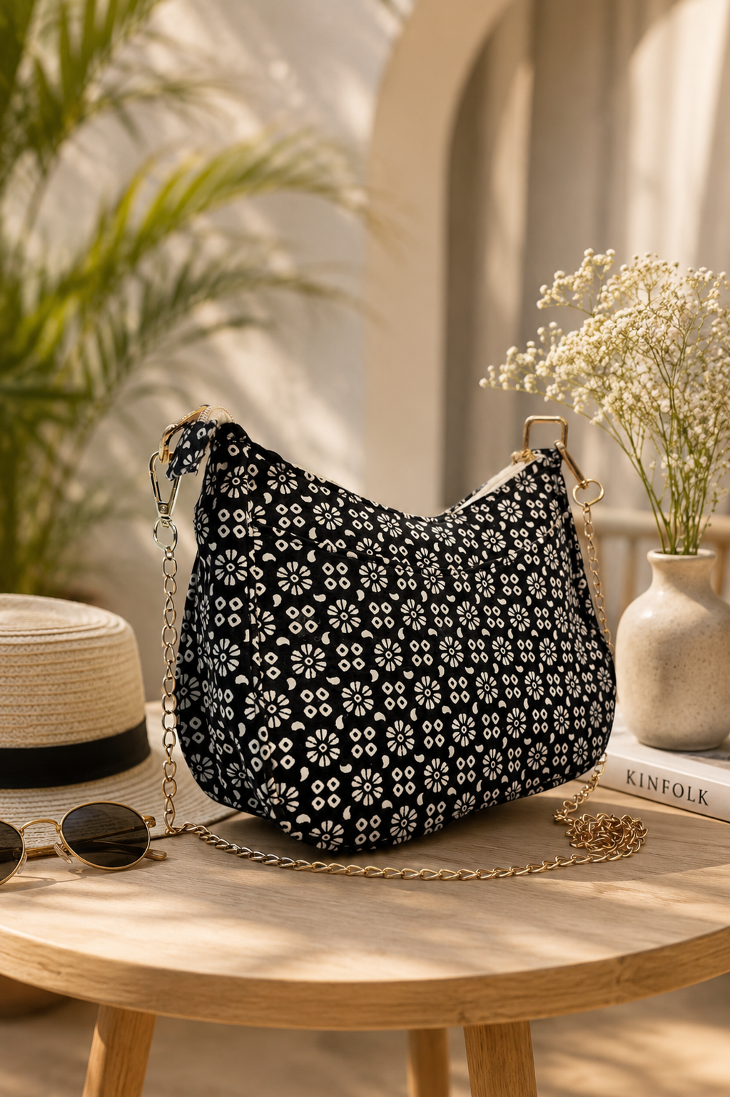 A black crescent-shaped shoulder bag with a white floral pattern, styled on a round wooden table with a straw hat, sunglasses, a book, and a vase of baby's breath in a sunlit indoor setting.