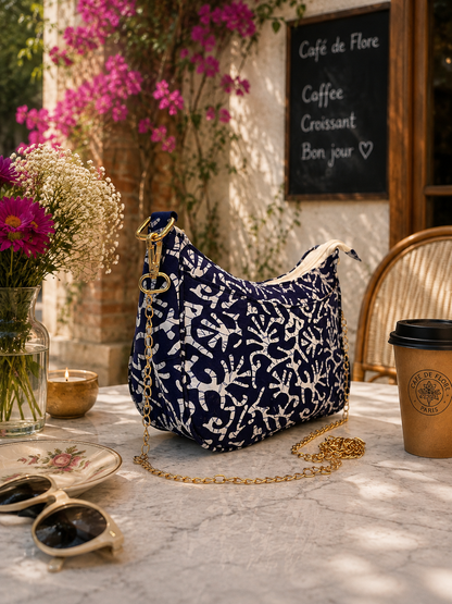 A navy blue and white patterned shoulder bag with a gold chain strap sits on a marble table in a cafe setting, surrounded by flowers, sunglasses, a coffee cup, and a chalkboard sign.