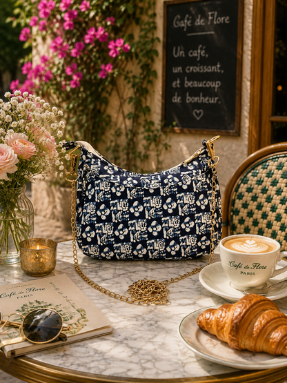 A navy blue and white patterned shoulder bag with a gold chain strap sits on a marble cafe table surrounded by a cup of latte, a croissant, flowers, and a chalkboard sign reading Cafe de Flore.
