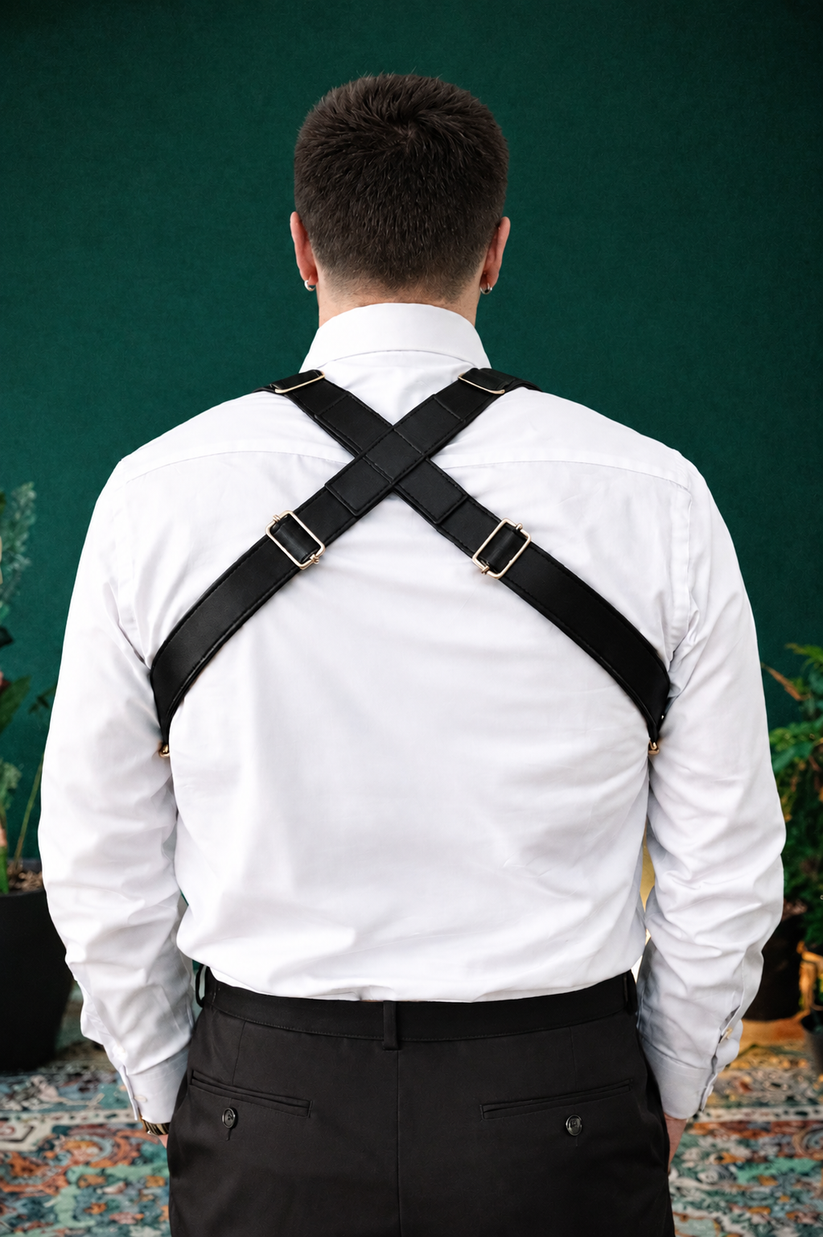 A man viewed from behind wearing black cross-back suspenders with silver metal buckles over a white dress shirt and dark trousers, standing against a dark green wall.