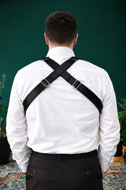 A man viewed from behind wearing black cross-back suspenders with silver metal buckles over a white dress shirt and dark trousers, standing against a dark green wall.