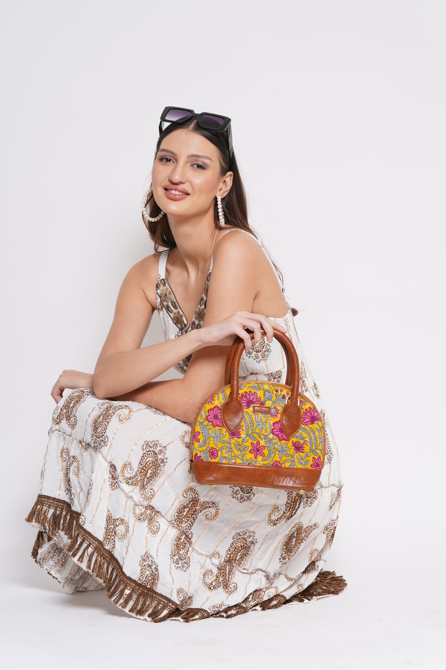 A woman wearing a white tiered dress with brown paisley patterns and holding a colorful floral handbag, posed against a plain white background.