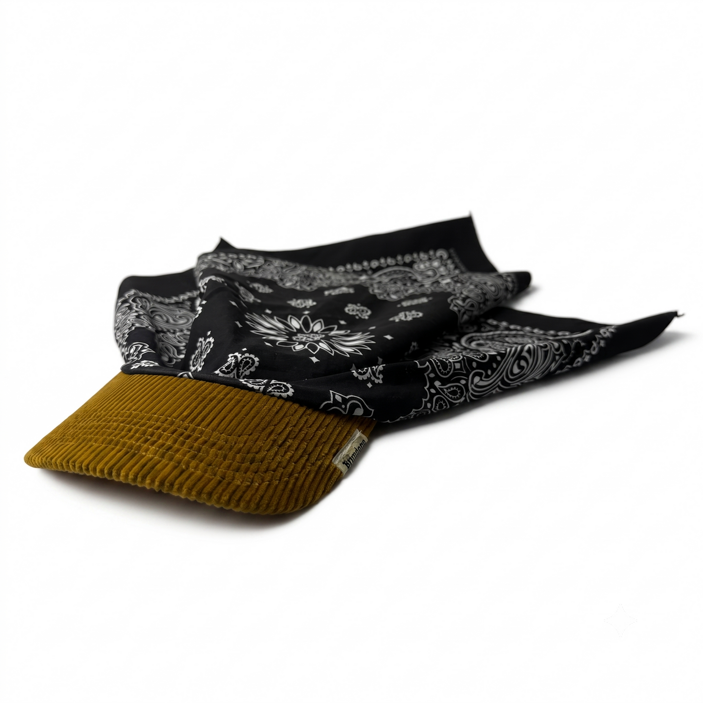 A black bandana with a white paisley pattern draped over the brim of a mustard yellow ribbed cap, shown on a clean white background.