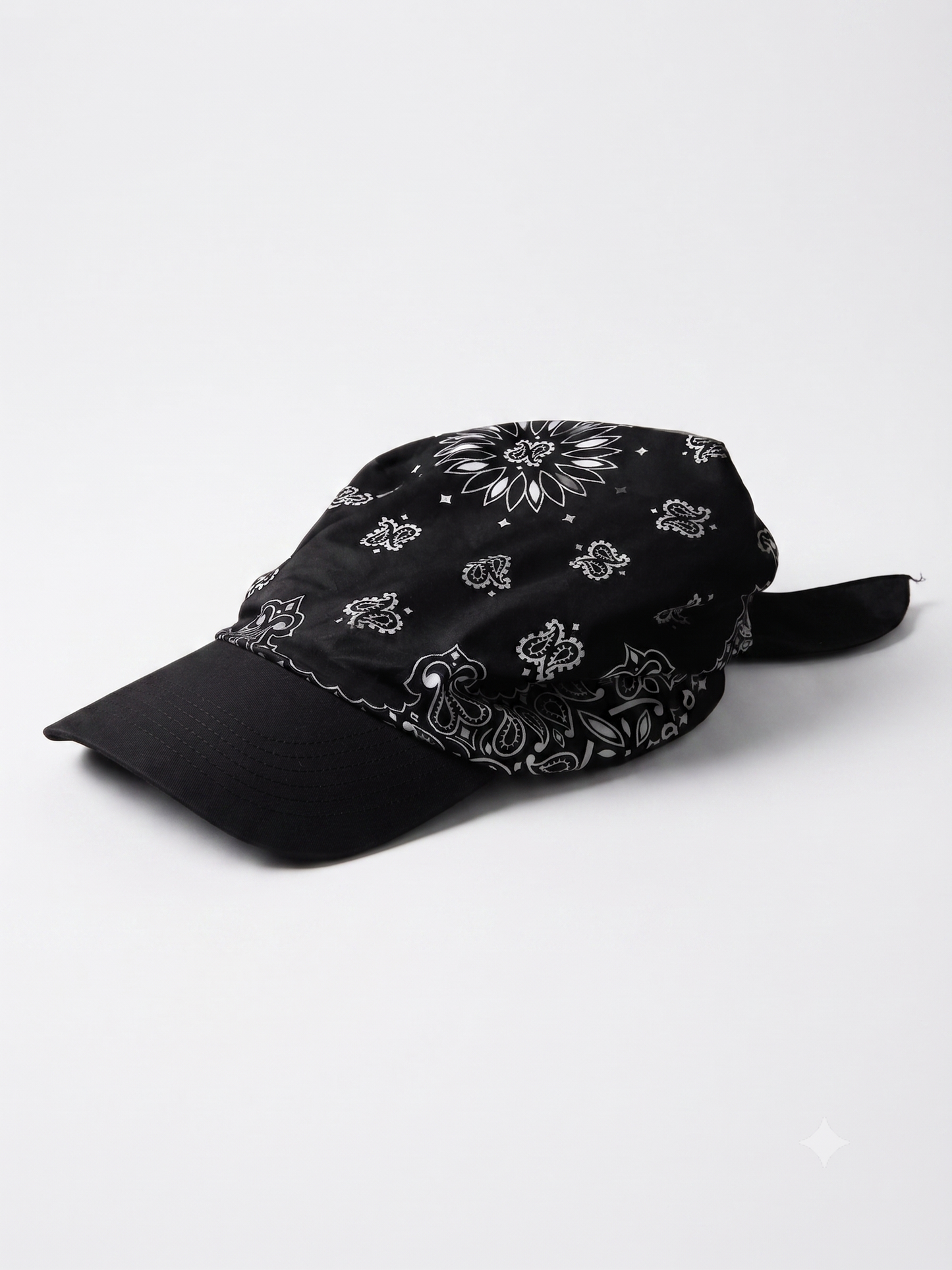 A black baseball cap with a white paisley bandana pattern on the crown and brim, featuring a black fabric chin strap, photographed on a light gray background.