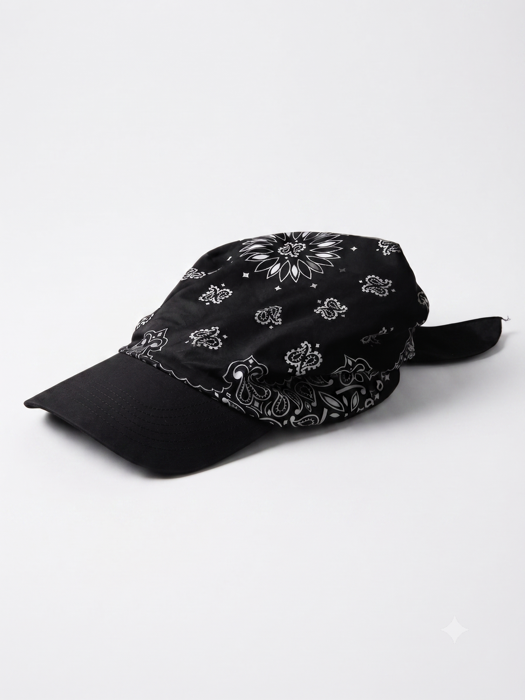 A black baseball cap with a white paisley bandana pattern on the crown and brim, featuring a black fabric chin strap, photographed on a light gray background.