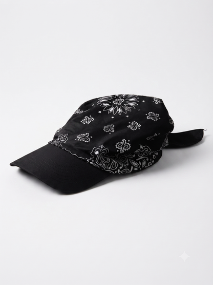 A black baseball cap with a white paisley bandana pattern on the crown and brim, featuring a black fabric chin strap, photographed on a light gray background.