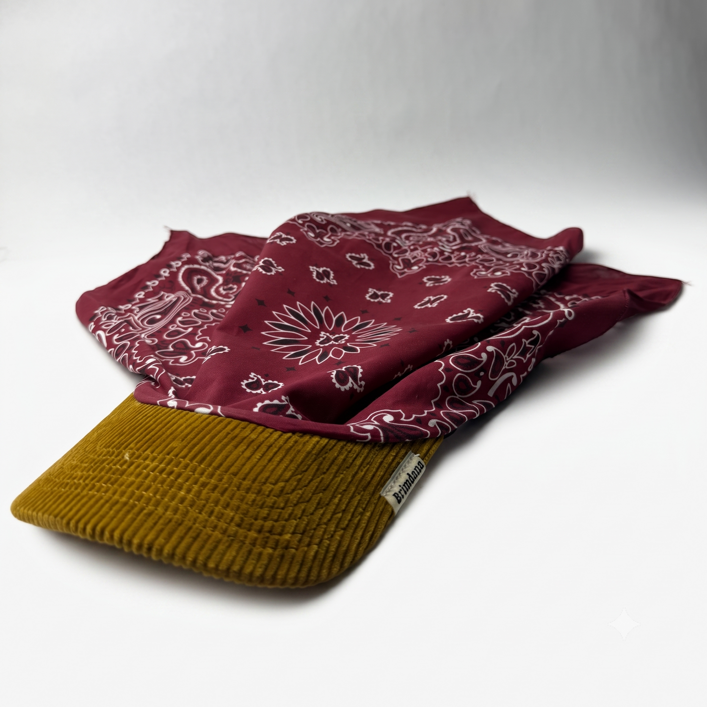 A burgundy bandana with a white paisley pattern draped over the brim of a mustard yellow corduroy cap, set against a plain light gray background.