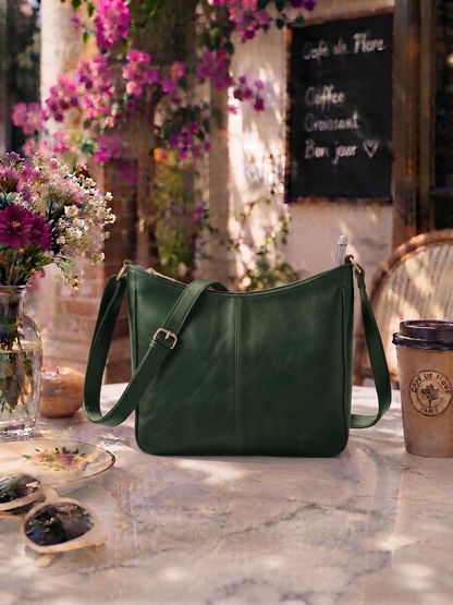 A dark green leather shoulder bag with gold-tone hardware sits on a stone table in a cafe setting, surrounded by flowers, sunglasses, a coffee cup, and a chalkboard menu.