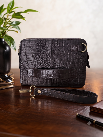 A dark brown crocodile-embossed leather crossbody bag with a matching wristlet strap, displayed on a polished wooden table next to a potted plant.