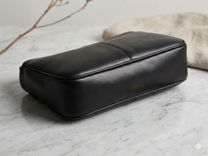 A black leather rectangular box or case with rounded corners, photographed on a light marble surface with a soft beige fabric prop in the background.