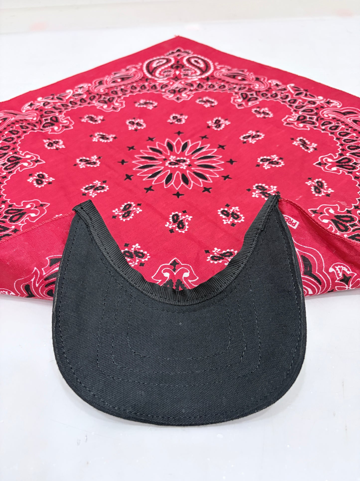 A black curved visor or hat brim placed in front of a folded red bandana with black paisley-style patterns on a white surface.