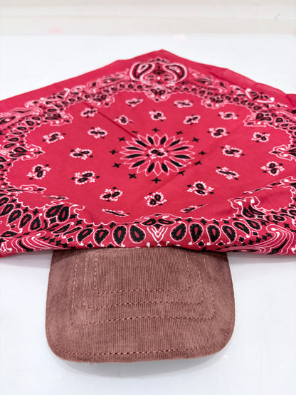 A red bandana with a black and white paisley pattern draped over the brim of a brown corduroy baseball cap, set against a plain white background.