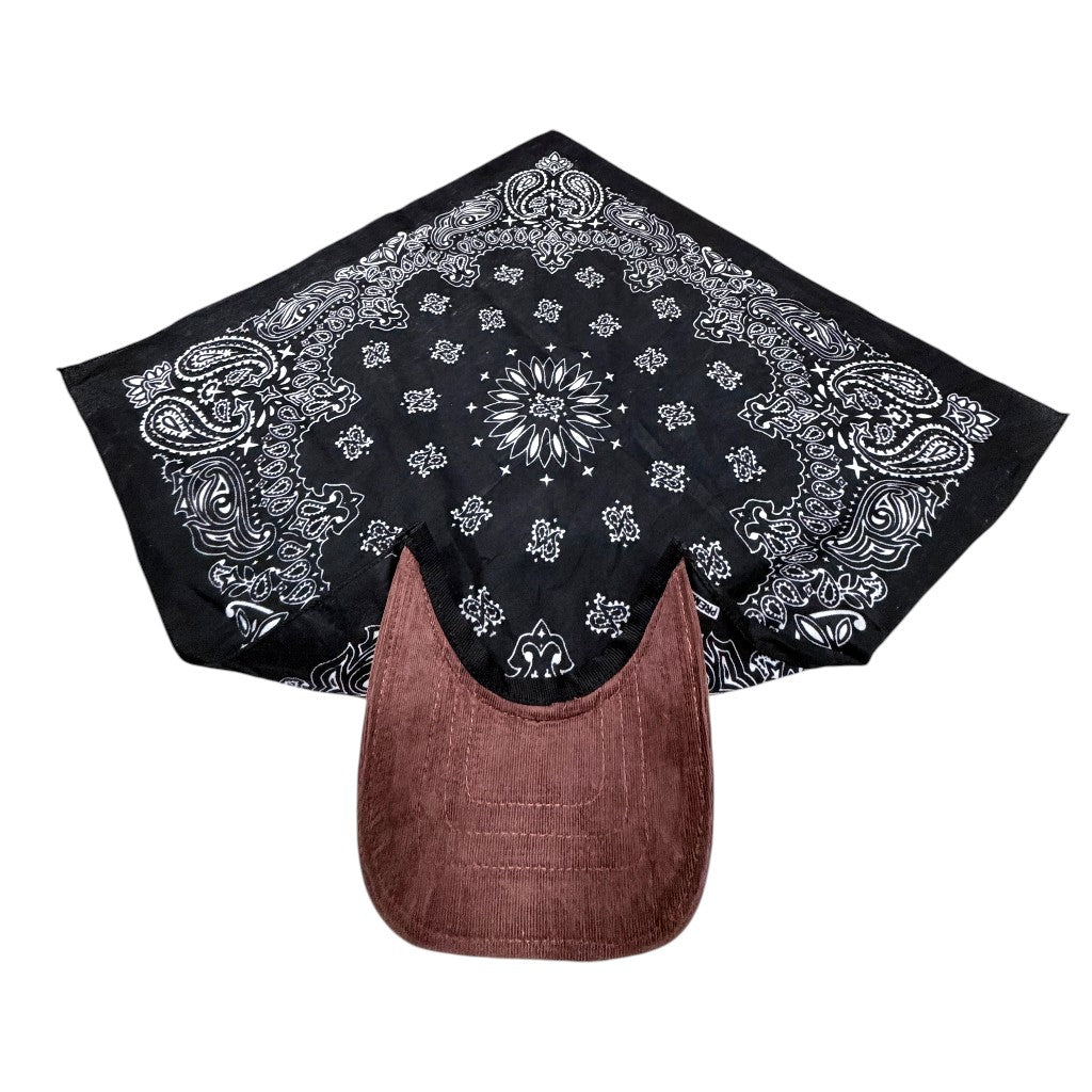 A black bandana with a white paisley and floral pattern, draped over a brown baseball cap on a plain white background.